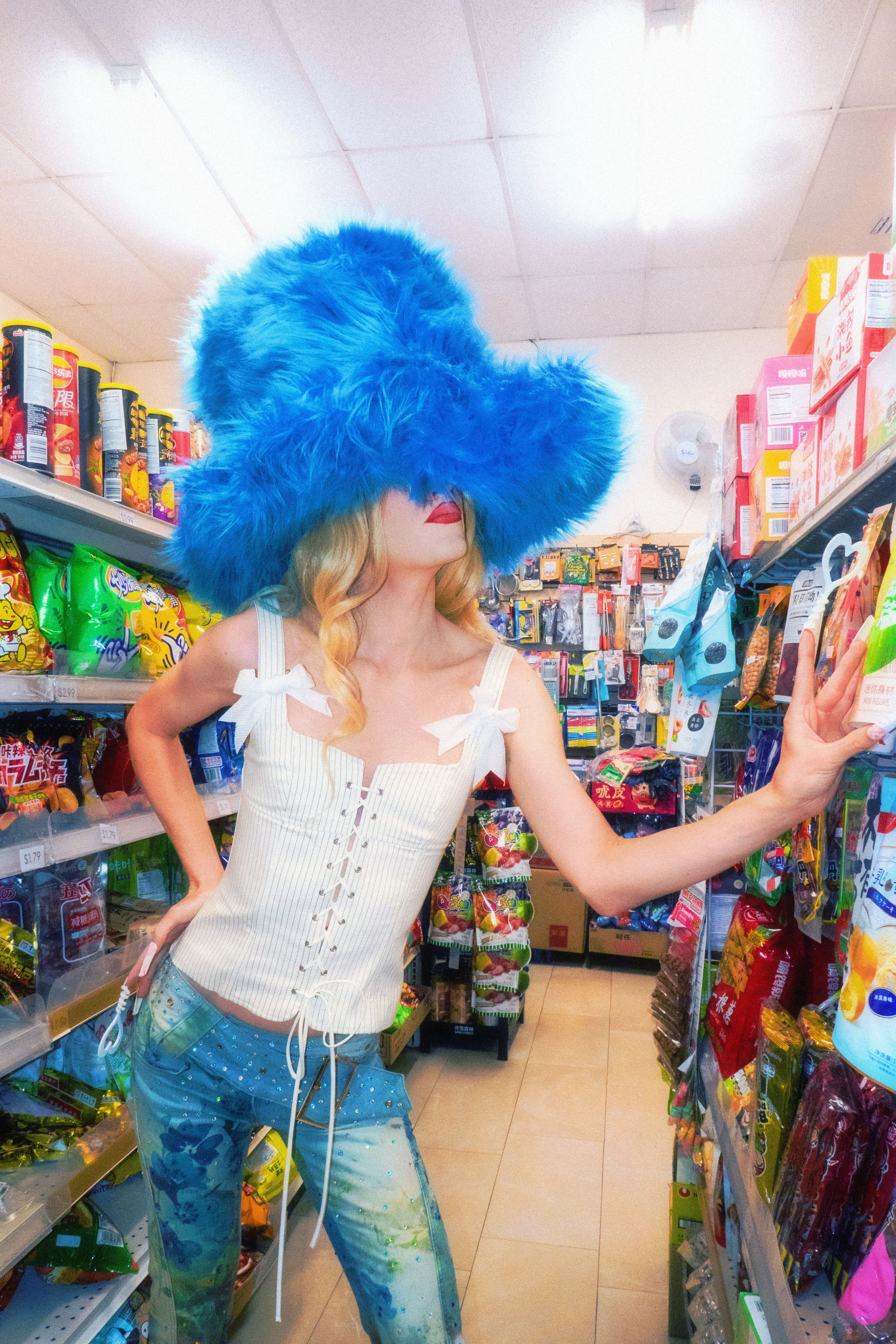 Jesse in y2k inspired drag reaching for candy with an oversized blue hat draped over their face in a colorful conveince store.